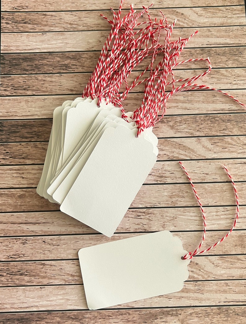 May include: A stack of white gift tags with scalloped edges and red and white baker's twine. The tags are on a woodgrain background.