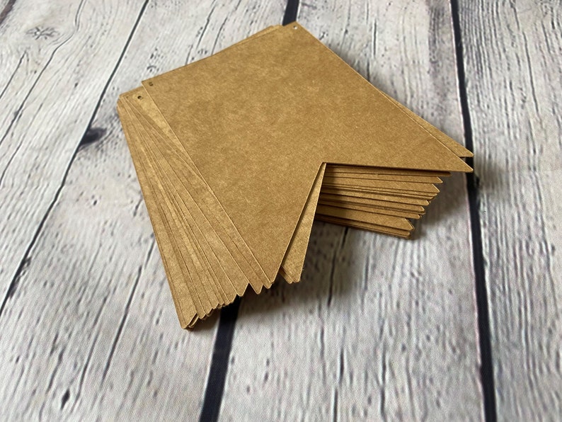 Puede incluir: Una pila de banderas de papel marr&oacute;n con agujeros perforados en la parte superior para colgar. Las banderas est&aacute;n dobladas por la mitad y apiladas una encima de la otra.