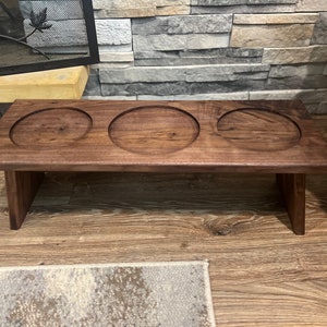 May include: A dark brown wooden serving tray with three circular cutouts. The tray is rectangular with short legs, designed to hold items. The wood grain is visible, and the tray sits on a wooden floor.