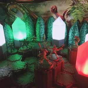Puede incluir: Una escena de juego de mesa con tres fuentes de luz de cristal brillantes. Los cristales son blancos, verdes y rojos. Los cristales están sobre una base y están rodeados de figuras en miniatura y terreno.