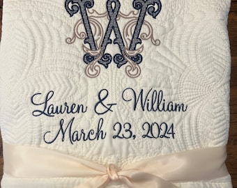 May include: A white quilted throw blanket with a navy blue and brown embroidered monogram "W" and the text "Lauren & William March 23, 2024". The blanket is tied with a cream-colored satin ribbon.