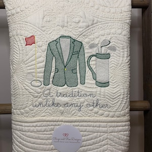 May include: A white quilted throw blanket with green embroidered images of a golf bag, golf clubs, a golfer's jacket, and a golf flag. The text "A tradition unlike any other" is embroidered below the images. The text "Chugs and Ree Designs Home Decor with Personality!" is embroidered in a circle at the bottom of the blanket.