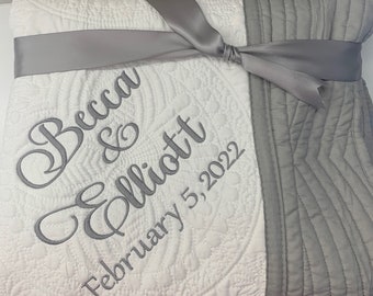 May include: A white and gray quilted blanket with a silver ribbon tied around it. The blanket has the names "Becca & Elliott" and the date "February 5, 2022" embroidered on it.