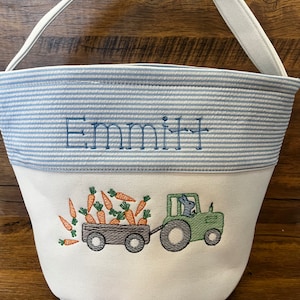 May include: A white canvas Easter basket with blue and white striped fabric lining. The basket has a white embroidered design of a bunny driving a green tractor pulling a wagon full of carrots. The basket has the name "Emmitt" embroidered on the front.