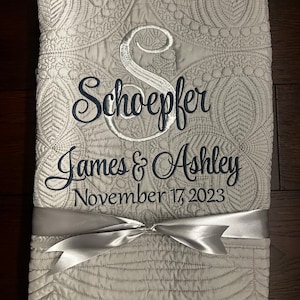 May include: A white quilted blanket with a silver satin ribbon tied in a bow. The blanket is embroidered with the text "Schoeffer James & Ashley November 17, 2023".