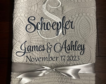 May include: A white quilted blanket with a silver satin ribbon tied in a bow. The blanket is embroidered with the text "Schoeffer James & Ashley November 17, 2023".