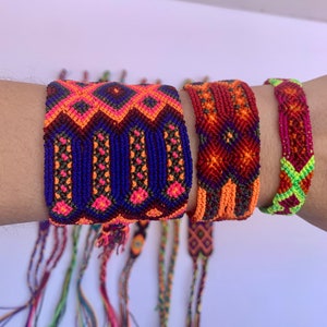 May include: A set of colorful friendship bracelets with geometric patterns in shades of orange, red, green, blue, and pink. The bracelets are made with woven thread and have tassels.