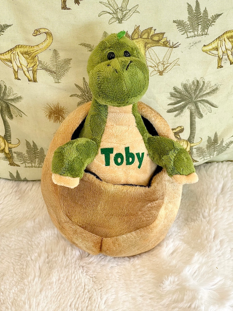 May include: Green plush dinosaur toy with a brown shell, sitting on a white fluffy surface. The dinosaur has a name tag that says "Toby".