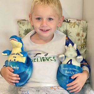 May include: A young boy is holding two blue plush dinosaur toys. The dinosaur on the left has the text "Look after me" and "Benjy" on it. The dinosaur on the right has the text "Happy birthday Benjy" and the number "4" on it. The boy is wearing a white shirt with the text "Believes" on it and grey pants with stars on them.