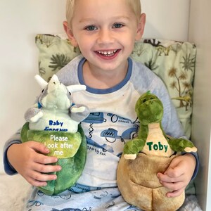 May include: A young boy is holding two plush dinosaur toys. The green dinosaur says "Toby" and the other dinosaur says "Baby dinosaur Please look after me".