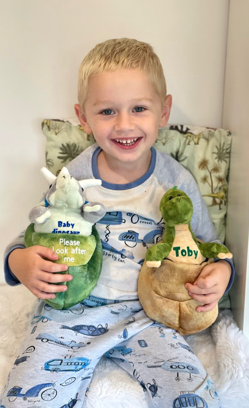 May include: A young boy is holding two plush dinosaur toys. The green dinosaur says "Toby" and the other dinosaur says "Baby dinosaur Please look after me".
