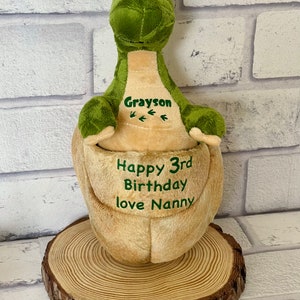 May include: Green plush dinosaur with a brown belly. The dinosaur is sitting on a wooden slice. The dinosaur has the name Grayson embroidered on its chest and the text "Happy 3rd Birthday love Nanny" embroidered on its belly.