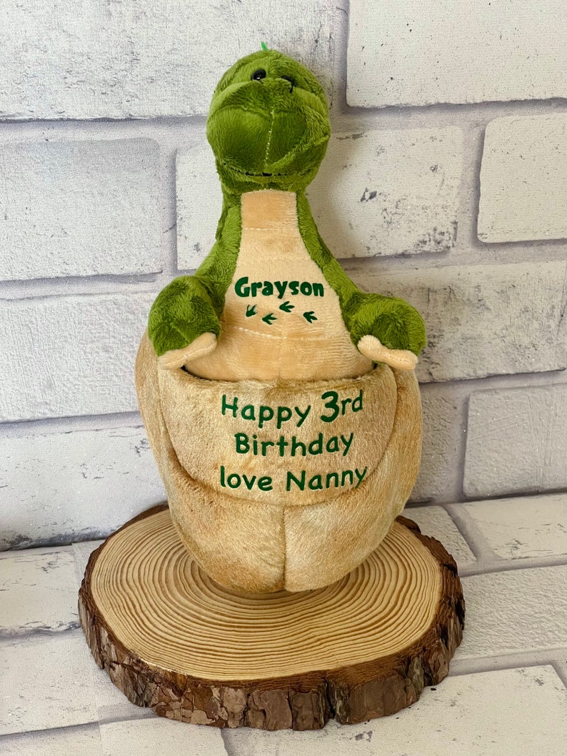 May include: Green plush dinosaur with a brown belly. The dinosaur is sitting on a wooden slice. The dinosaur has the name Grayson embroidered on its chest and the text "Happy 3rd Birthday love Nanny" embroidered on its belly.