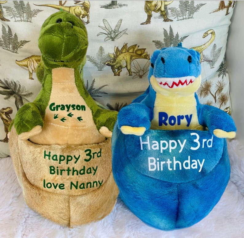 May include: Two plush dinosaur toys, one green and one blue, with personalized embroidery. The green dinosaur says "Grayson Happy 3rd Birthday love Nanny" and the blue dinosaur says "Rory Happy 3rd Birthday".