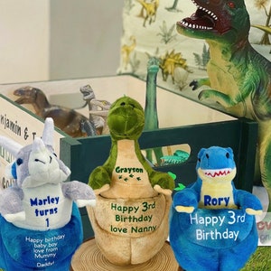 May include: Three plush dinosaur toys with personalized embroidered names and birthday messages. The dinosaurs are green, blue, and gray. The green dinosaur says "Grayson Happy 3rd Birthday love Nanny". The blue dinosaur says "Rory Happy 3rd Birthday". The gray dinosaur says "Marley turns 1 Happy birthday baby boy love from Mummy Daddy!"