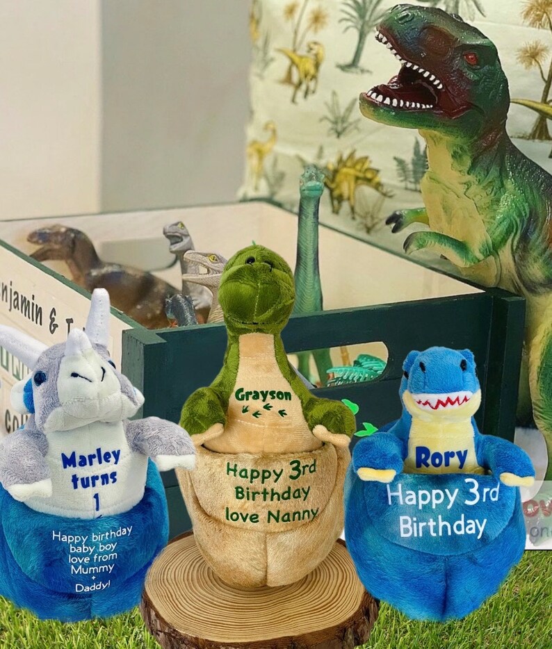 May include: Three plush dinosaur toys with personalized embroidered names and birthday messages. The dinosaurs are green, blue, and gray. The green dinosaur says "Grayson Happy 3rd Birthday love Nanny". The blue dinosaur says "Rory Happy 3rd Birthday". The gray dinosaur says "Marley turns 1 Happy birthday baby boy love from Mummy Daddy!"