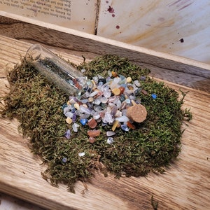 Könnte beinhalten: Ein klares Glasfläschchen gefüllt mit kleinen, bunten Edelsteinen, die teilweise auf ein Bett aus grünem Moos verschüttet wurden. Ein kleiner Korken liegt daneben. Die Anordnung liegt auf einem Holztablett.
