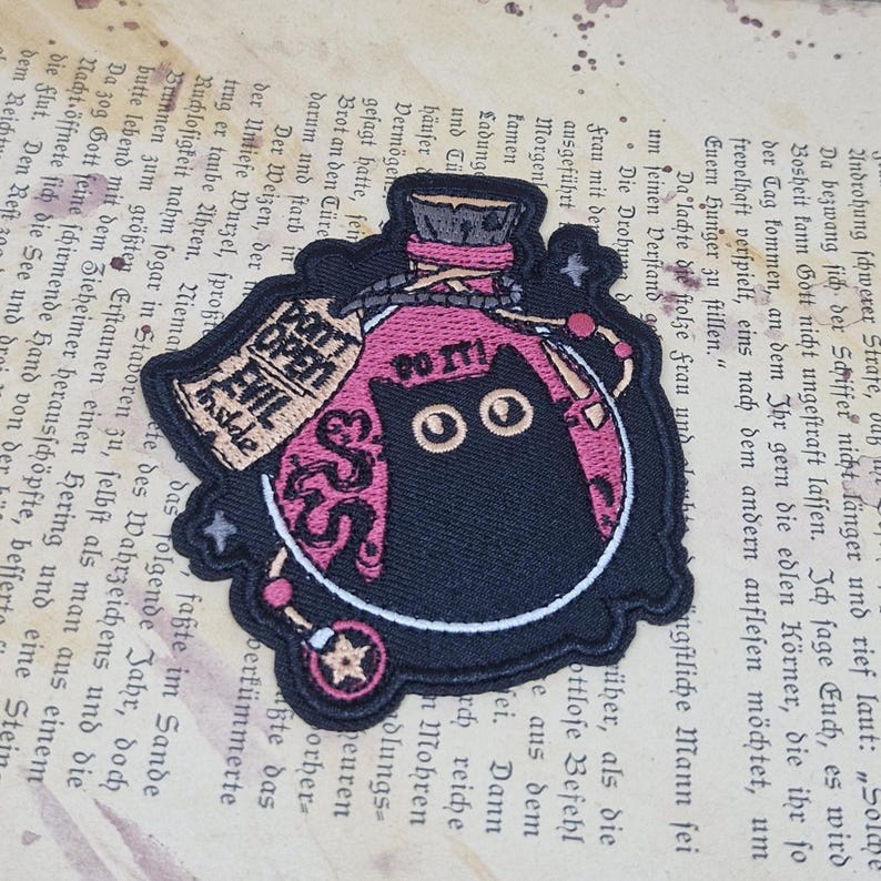May include: Embroidered patch featuring a black cat inside a pink potion bottle. The patch includes the text "DO IT!" and "DON'T OPEN EVIL INSIDE". Decorative beads and stars surround the bottle, creating a whimsical design.