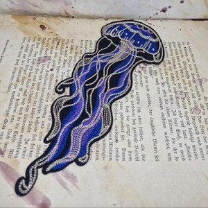 May include: Embroidered jellyfish patch in shades of blue, black, and silver. The design features intricate details and a flowing, stylized form. This patch is suitable for jackets, bags, or other fabric items.