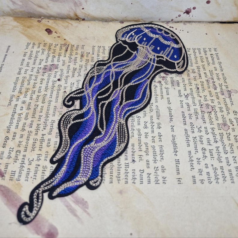 May include: Embroidered jellyfish patch in shades of blue, black, and silver. The design features intricate details and a flowing, stylized form. This patch is suitable for jackets, bags, or other fabric items.
