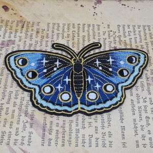 May include: Embroidered butterfly patch with a celestial design. The butterfly features a blue body, black wings with white stars, and a gold outline. The patch is on a vintage paper background.