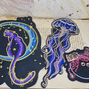 May include: Embroidered patches featuring a cat on a crescent moon, a jellyfish, and a potion bottle with the text "DO IT!" and "EVIL Inside". The patches are black with purple, blue, and gold accents. The patches are on a vintage book page.