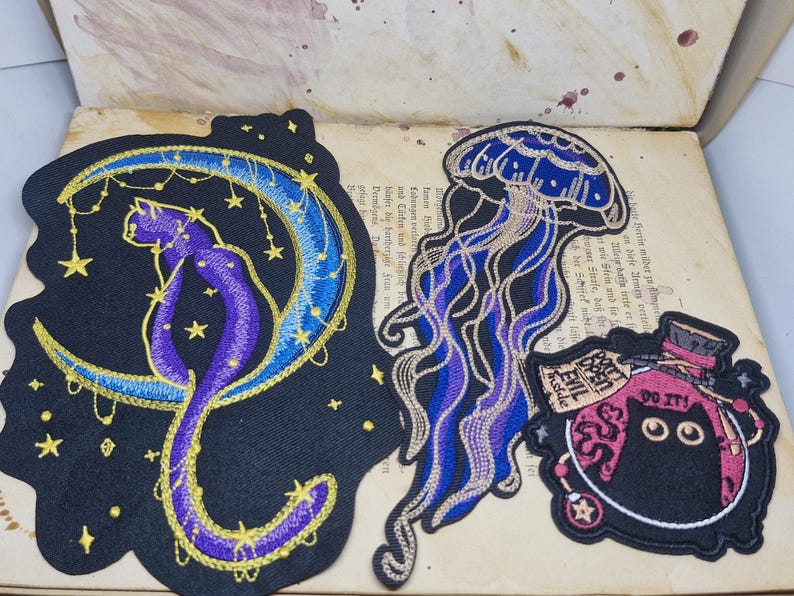 May include: Embroidered patches featuring a cat on a crescent moon, a jellyfish, and a potion bottle with the text "DO IT!" and "EVIL Inside". The patches are black with purple, blue, and gold accents. The patches are on a vintage book page.
