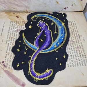 May include: Embroidered patch featuring a purple cat sitting on a crescent moon. The moon is blue with gold embroidery, and the cat has gold accents. Stars dangle from the moon, all on a black background.