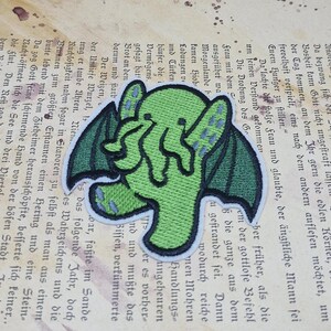 May include: Green Cthulhu patch with black outline and wings, set against a vintage book page background. The patch features a stylized design of the mythical creature. Ideal for jackets, bags, or other fabric items.