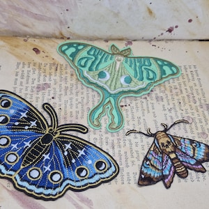 Könnte beinhalten: Drei gestickte Aufnäher mit Schmetterlingen und einer Motte in Blau-, Grün- und Brauntönen. Sie liegen auf einer Vintage-Buchseite mit deutschem Text. Der blaue Schmetterling hat weiße Sterne.