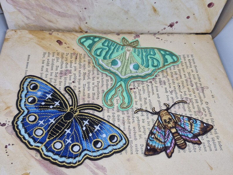 May include: Three embroidered butterfly and moth patches in shades of blue, green, and brown. The patches are on a vintage book page with German text. The blue butterfly has white star details.