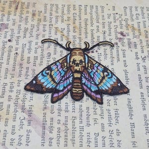 May include: Embroidered moth patch with a skull design on a vintage book page. The moth features intricate details with brown, blue, and purple wings. This patch is ideal for adding a unique touch to clothing or accessories.