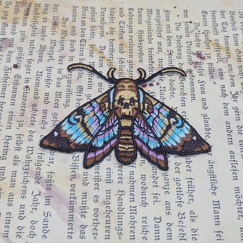 May include: Embroidered moth patch with a skull design on a vintage book page. The moth features intricate details with brown, blue, and purple wings. This patch is ideal for adding a unique touch to clothing or accessories.