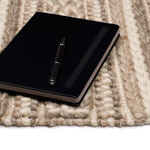 May include: A black leather-bound journal with a silver pen lying on top of a beige and white woven rug.