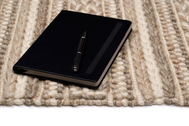 May include: A black leather-bound journal with a silver pen lying on top of a beige and white woven rug.