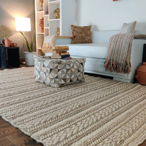 May include: A white and beige striped rug with a geometric patterned ottoman in the foreground. The ottoman is covered in a patchwork of light brown, gray, and white leather. A white sofa with a throw blanket and pillows is in the background.