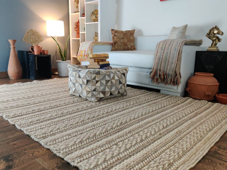 May include: A white and beige striped rug with a geometric patterned ottoman in the foreground. The ottoman is covered in a patchwork of light brown, gray, and white leather. A white sofa with a throw blanket and pillows is in the background.