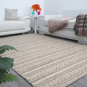 May include: A beige and white striped woven rug with a textured pattern. The rug is placed in a living room setting with white couches and a small table with a vase of orange flowers.