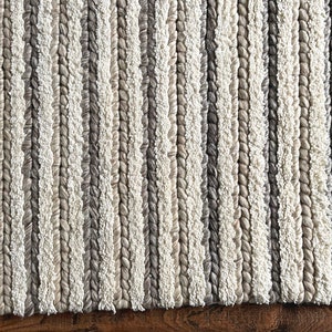May include: A close-up of a textured rug with vertical stripes in shades of white, gray, and beige. The rug has a braided appearance, with the stripes creating a pattern. The rug's border is a darker gray.
