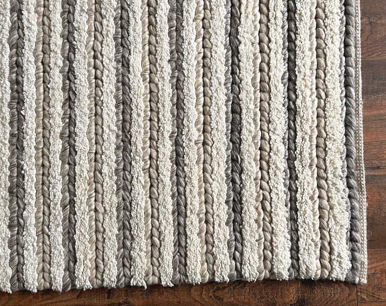 May include: A close-up of a textured rug with vertical stripes in shades of white, gray, and beige. The rug has a braided appearance, with the stripes creating a pattern. The rug's border is a darker gray.