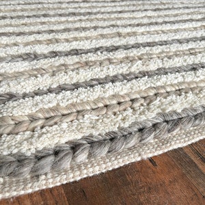 May include: A textured rug with horizontal stripes in shades of white, gray, and beige. The rug features braided and looped textures, with a label that reads "THE LOOMINAIRE". The rug is placed on a wooden surface.