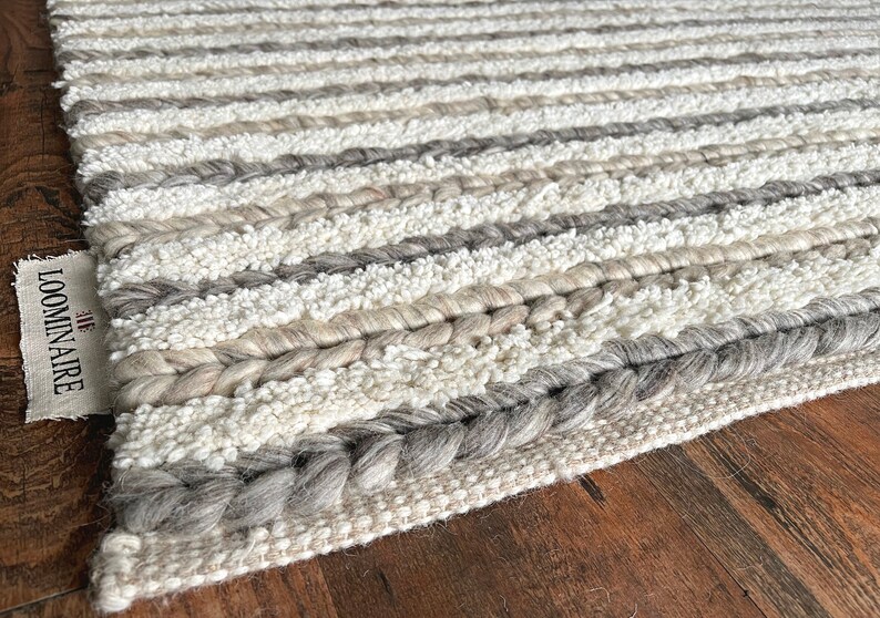 May include: A textured rug with horizontal stripes in shades of white, gray, and beige. The rug features braided and looped textures, with a label that reads "THE LOOMINAIRE". The rug is placed on a wooden surface.