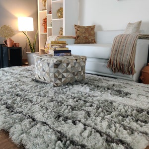 May include: A gray and white shag rug with a geometric patterned ottoman in the center of the room. The ottoman is made of leather and has a light brown, gray, and white color scheme.