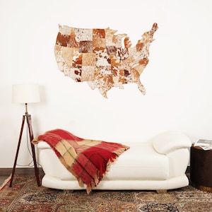 May include: A cowhide map of the United States hangs on a white wall. The map is made of various patches of brown, tan, and white cowhide. Below the map is a white chaise lounge with a red and tan plaid blanket.