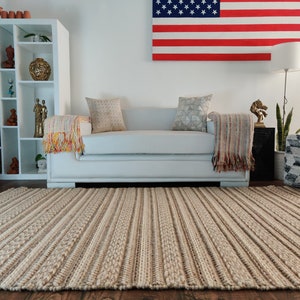 May include: A light beige striped rug with a woven texture. The rug is in a living room with a white sofa, a white bookshelf, and a decorative American flag on the wall.