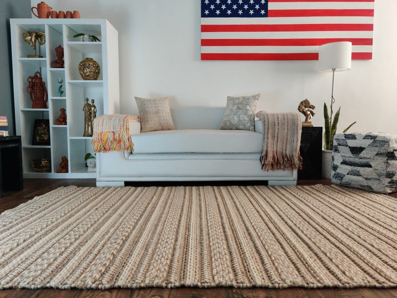 May include: A light beige striped rug with a woven texture. The rug is in a living room with a white sofa, a white bookshelf, and a decorative American flag on the wall.