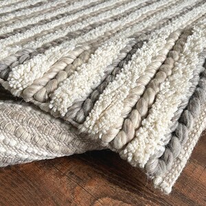 May include: Close-up of a textured rug with a mix of neutral colors. The rug features alternating stripes of braided and looped textures in shades of white, gray, and beige. The design creates a cozy and inviting feel, suitable for home decor.