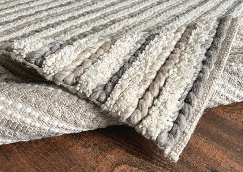 May include: Close-up of a textured rug with a mix of neutral colors. The rug features alternating stripes of braided and looped textures in shades of white, gray, and beige. The design creates a cozy and inviting feel, suitable for home decor.