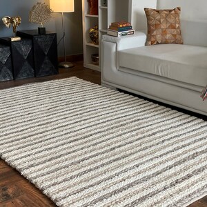 May include: A rectangular rug with a striped pattern in shades of white, beige, and gray. The rug is made of a textured material and is placed on a hardwood floor. The rug is a decorative element for a living room.