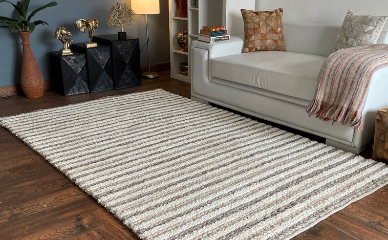 May include: A rectangular rug with a striped pattern in shades of white, beige, and gray. The rug is made of a textured material and is placed on a hardwood floor. The rug is a decorative element for a living room.